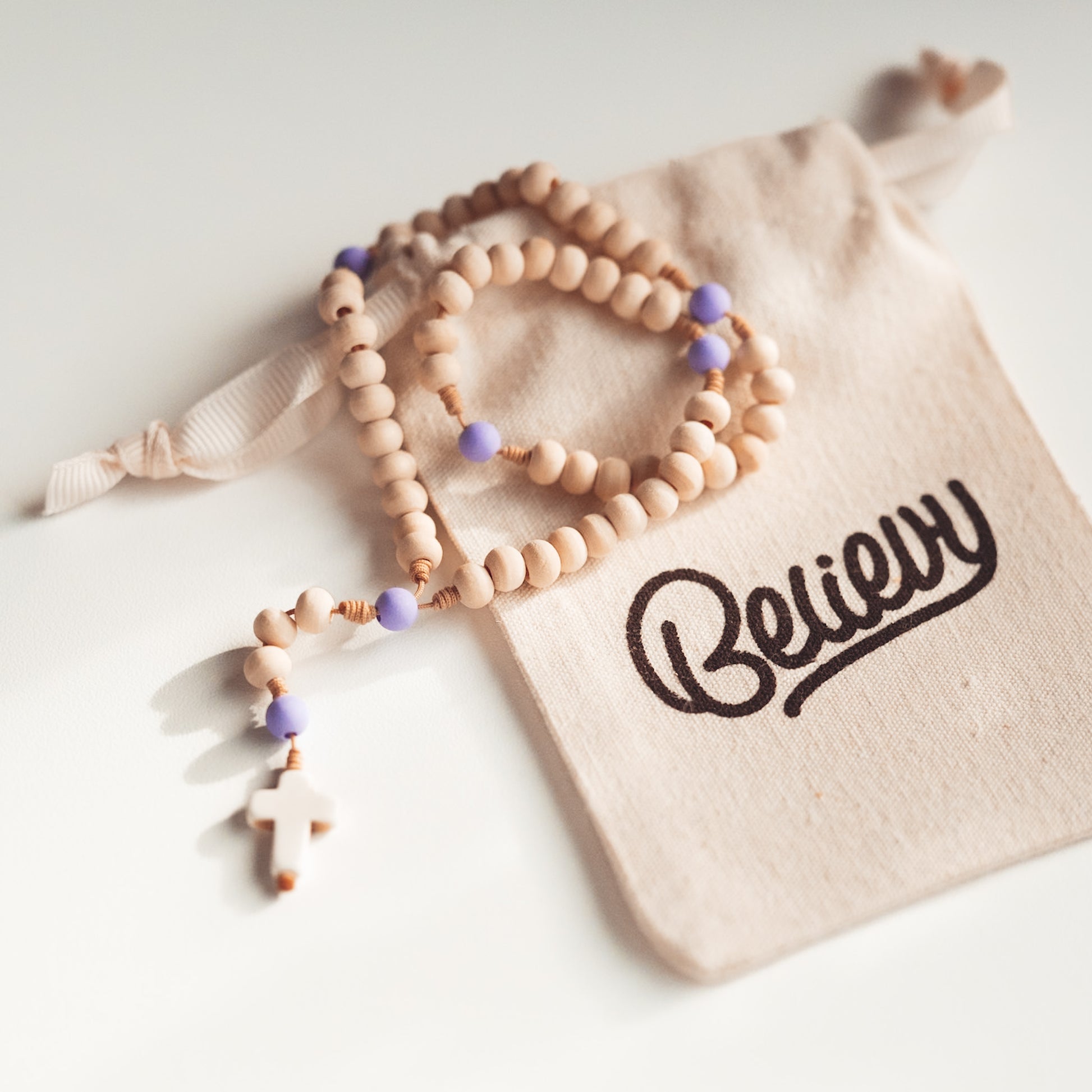 Drewniany różaniec z małym białym krzyżykiem i fioletowymi akcentami, sfotografowany w miękkim świetle na woreczku Believy.