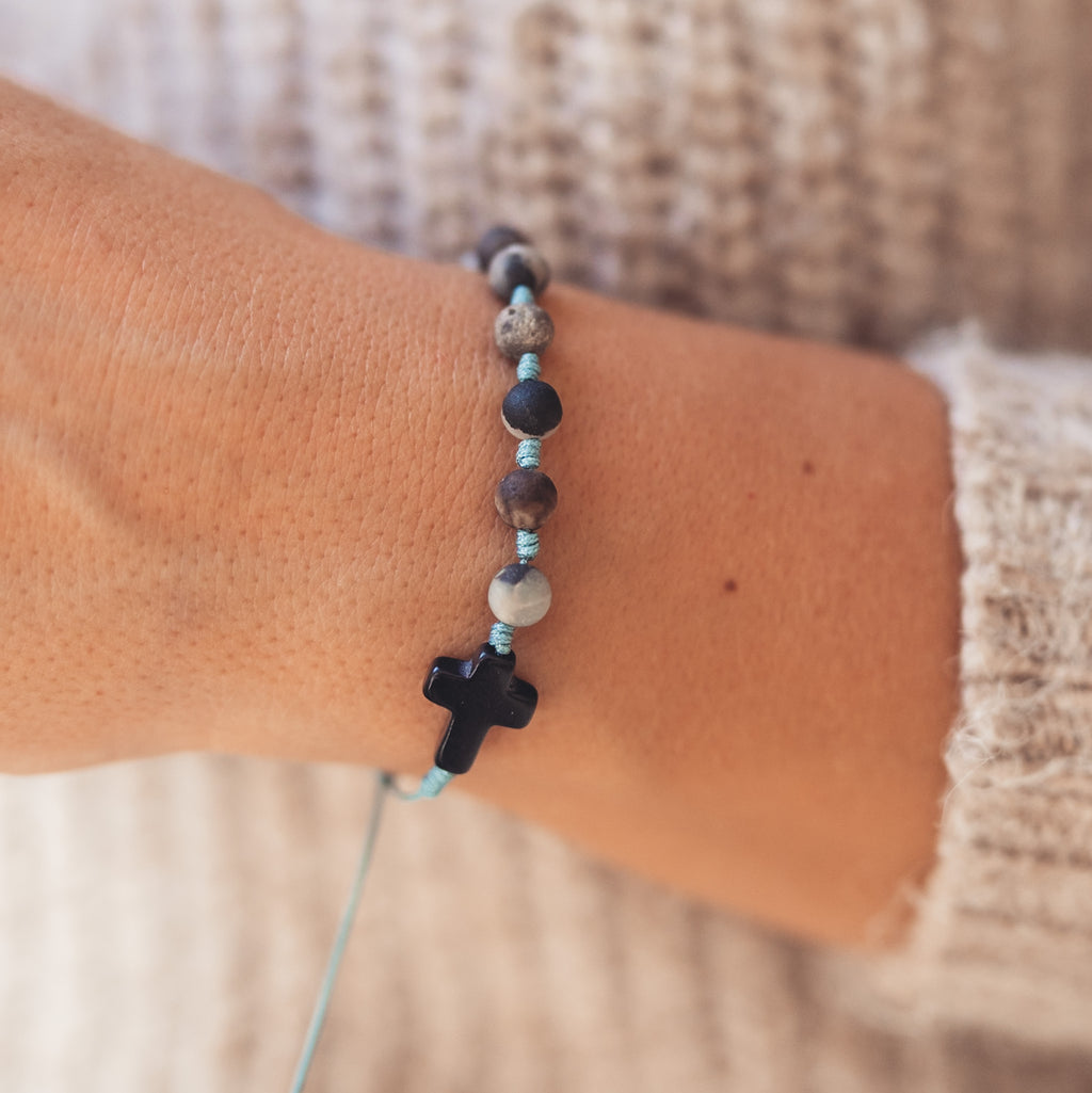 Zbliżenie na nadgarstek kobiety w beżowym swetrze, ozdobiony bransoletką (różańcem) z turkusowym sznurkiem, kamieniami w odcieniach szarości i czarnym krzyżykiem z kamienia.