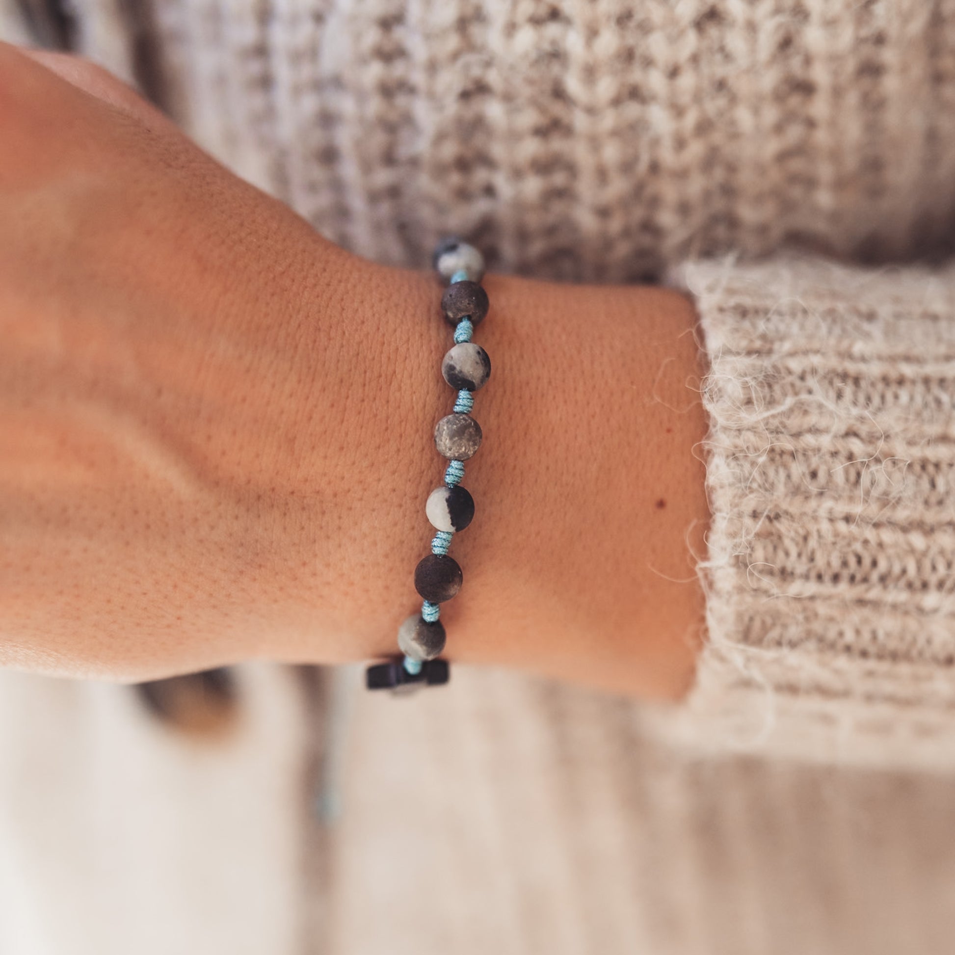 Różaniec z kamieni w tonacji brązowo-szarej, spleciona na błękitnym sznurku, widoczna na tle miękkiego, kremowego swetra.