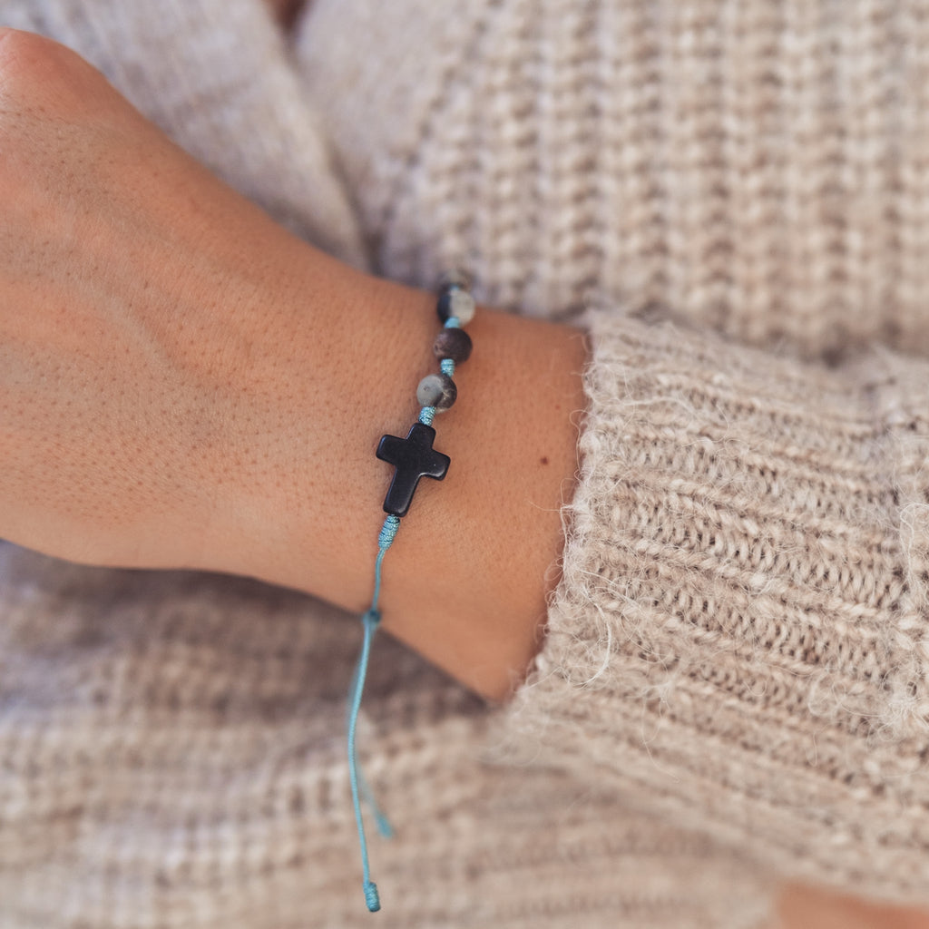 Ujęcie bransoletki - różańca z czarnym krzyżykiem i matowych kamieni w naturalnych barwach ziemi, na błękitnym sznurku, noszonej na nadgarstku kobiety w ciepłym swetrze.