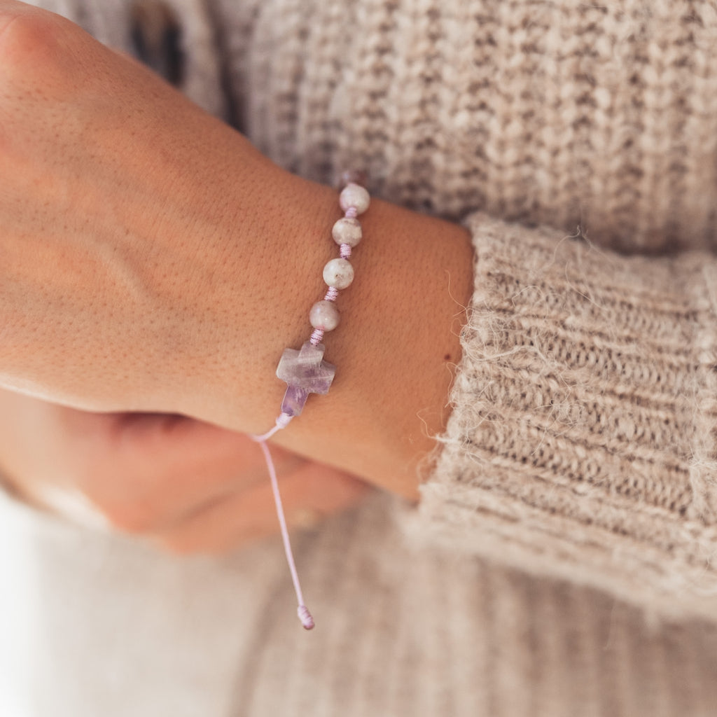 Zbliżenie na kobiecą dłoń ubraną w beżowy, dziergany sweter. Na nadgarstku widoczna delikatna bransoletka z lawendowym sznurkiem i kamiennym krzyżykiem w odcieniach fioletu.