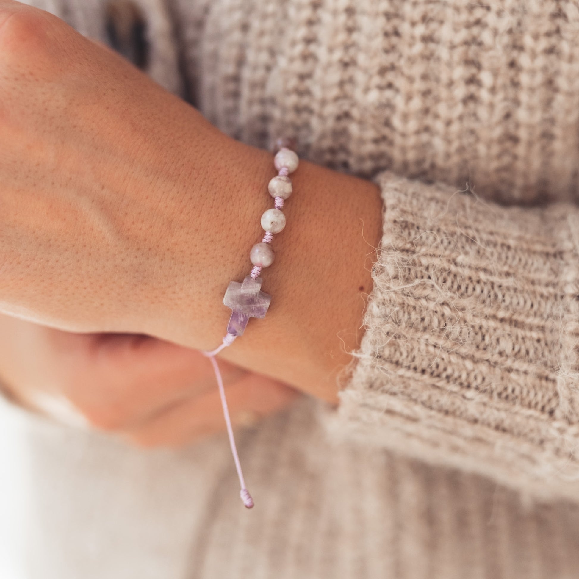 Zbliżenie na kobiecą dłoń ubraną w beżowy, dziergany sweter. Na nadgarstku widoczna delikatna bransoletka z lawendowym sznurkiem i kamiennym krzyżykiem w odcieniach fioletu.