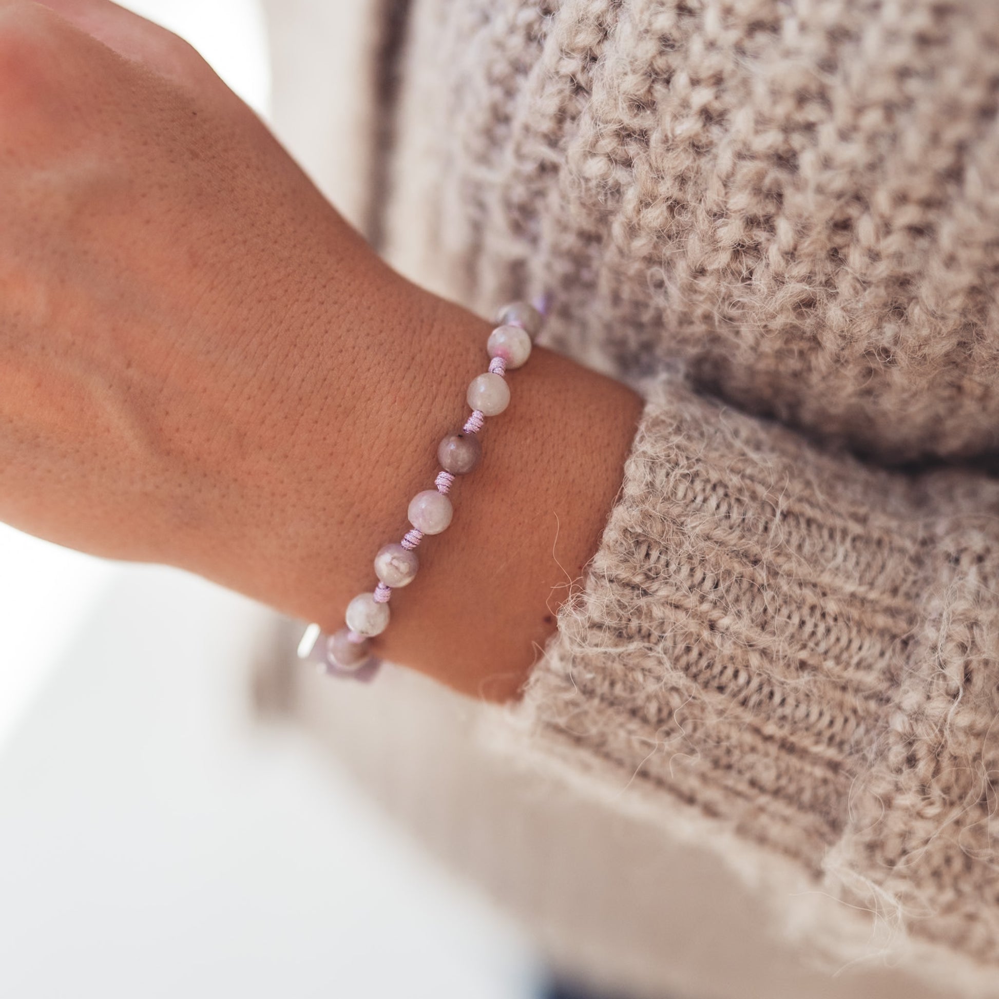 Bransoletka z jasnofioletowych kamieni na nadgarstku kobiety w beżowym swetrze, ujęcie z góry ukazujące subtelny połysk koralików.