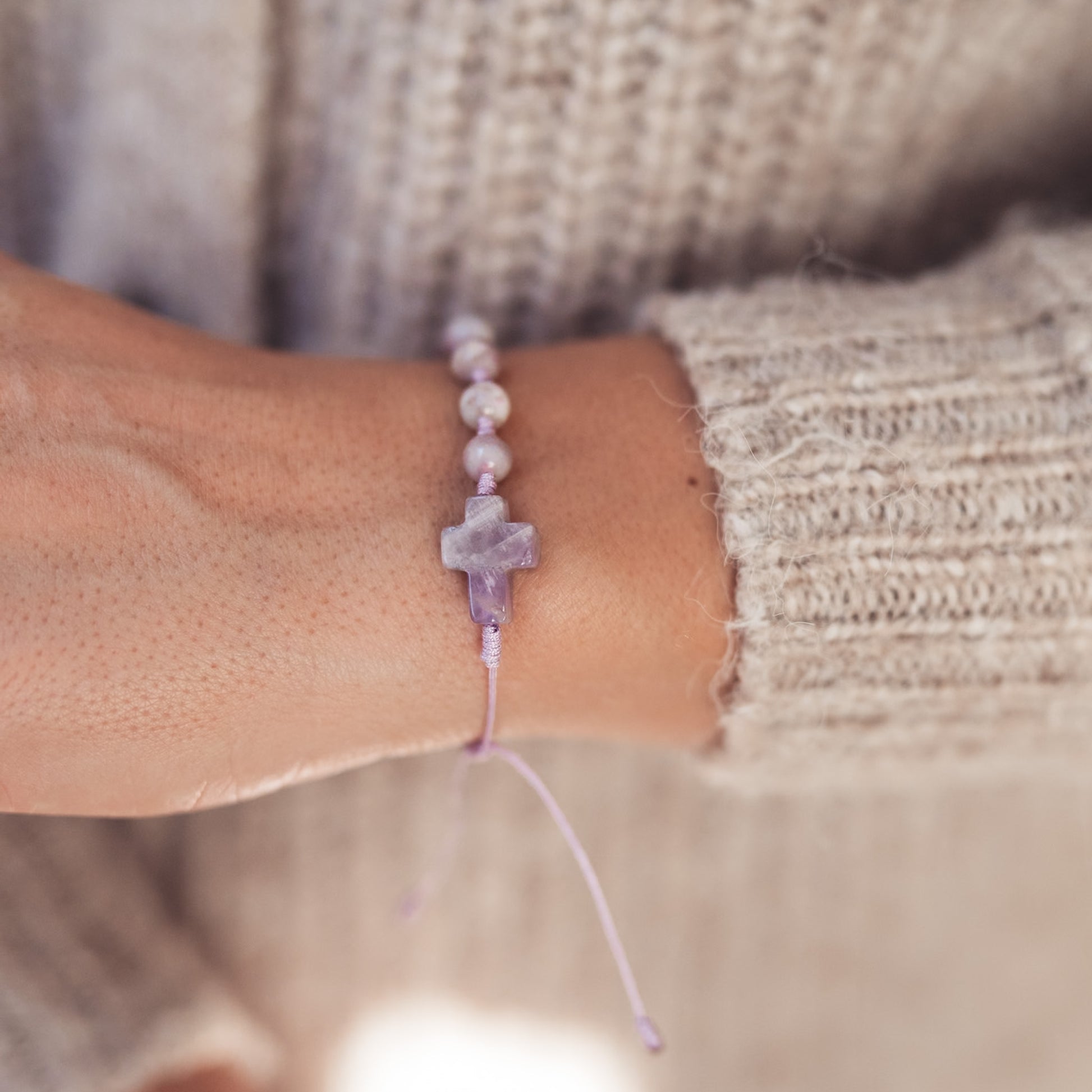 Ujęcie bransoletki z krzyżykiem z kamienia w odcieniach ametystu, splecionej na pastelowym sznurku, widoczne na tle miękkiego, kremowego swetra.