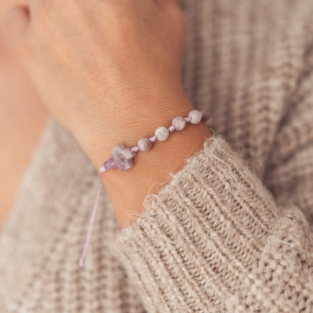 Bransoletka z kamiennym krzyżykiem i  koralikami w odcieniach różu i lawendy, na nadgarstku w miękkim beżowym swetrze, ujęcie z boku.