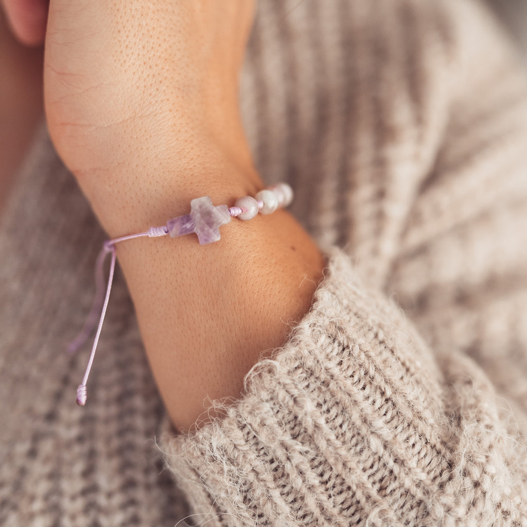 Zbliżenie na nadgarstek kobiety z bransoletką z krzyżykiem z fioletowego kamienia i pastelowych koralików, splecioną na delikatnym, liliowym sznurku.