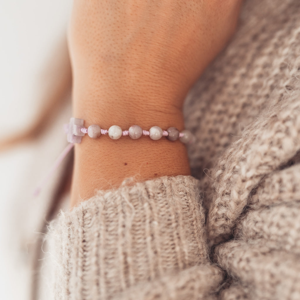 Widok bransoletki z drobnych, jasnych kamieni i krzyżyka z ametystu na nadgarstku kobiety w swetrze o grubym splocie, w miękkim, naturalnym świetle.