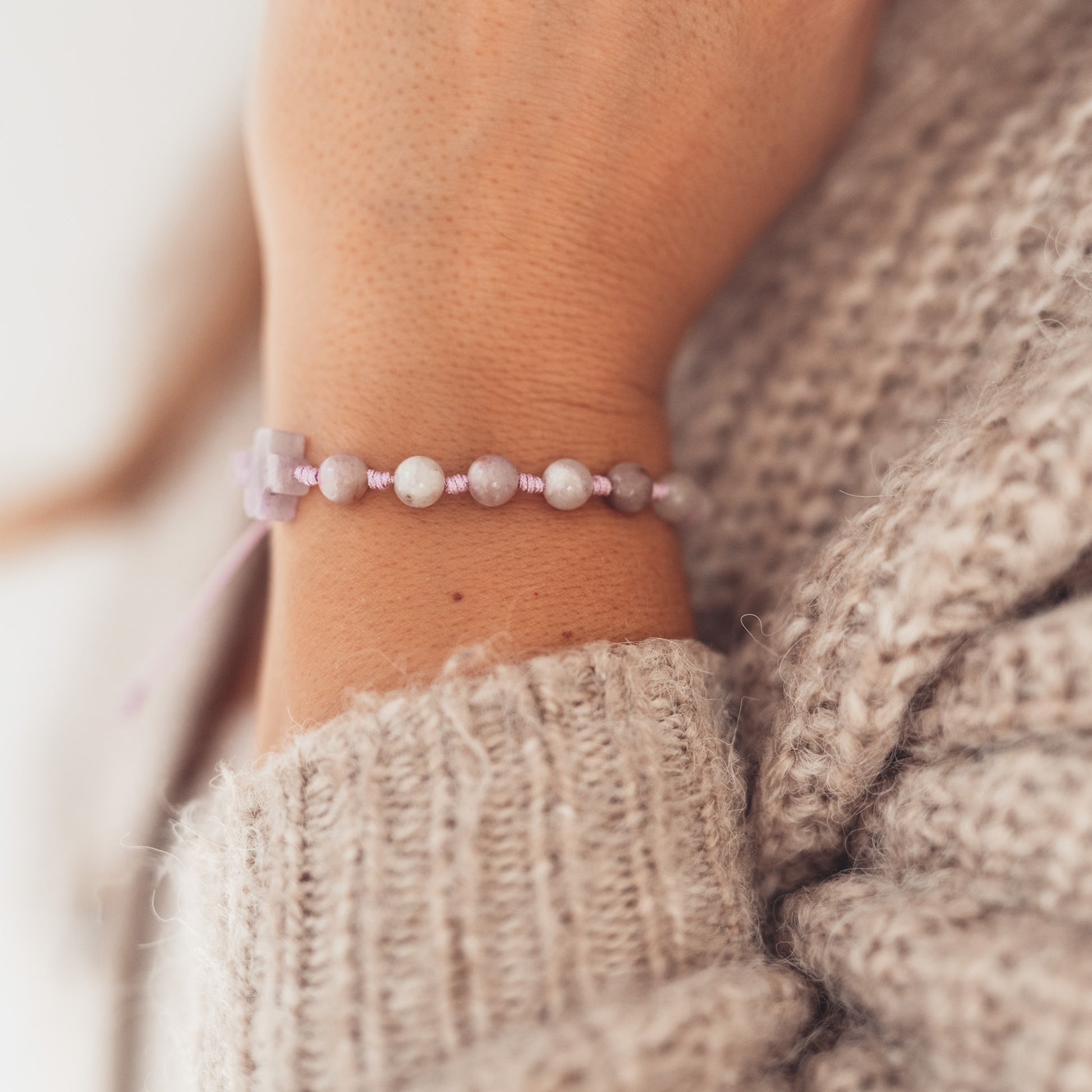 Widok bransoletki z drobnych, jasnych kamieni i krzyżyka z ametystu na nadgarstku kobiety w swetrze o grubym splocie, w miękkim, naturalnym świetle.