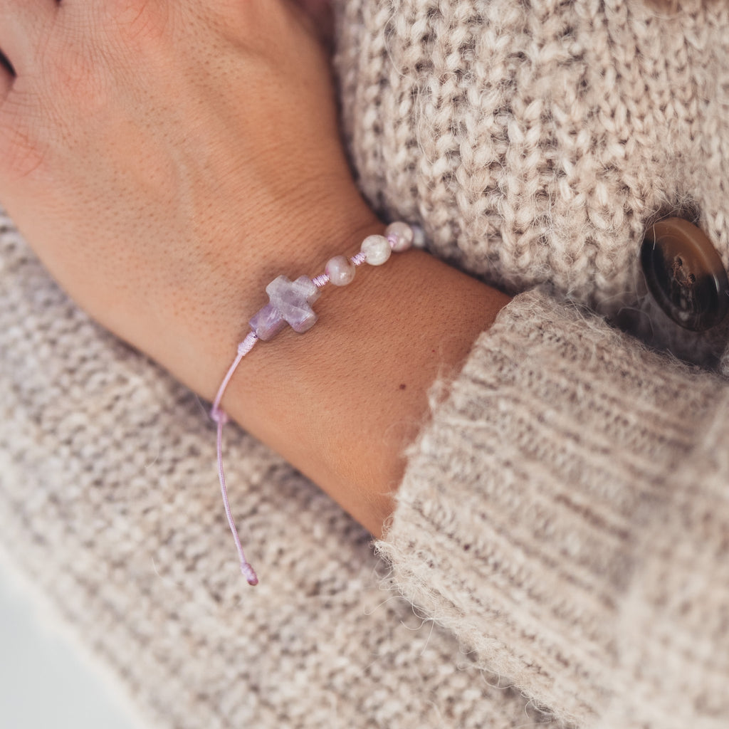 Widok bransoletki z drobnych, jasnych kamieni i krzyżyka z ametystu na nadgarstku kobiety w swetrze o grubym splocie, w miękkim, naturalnym świetle.