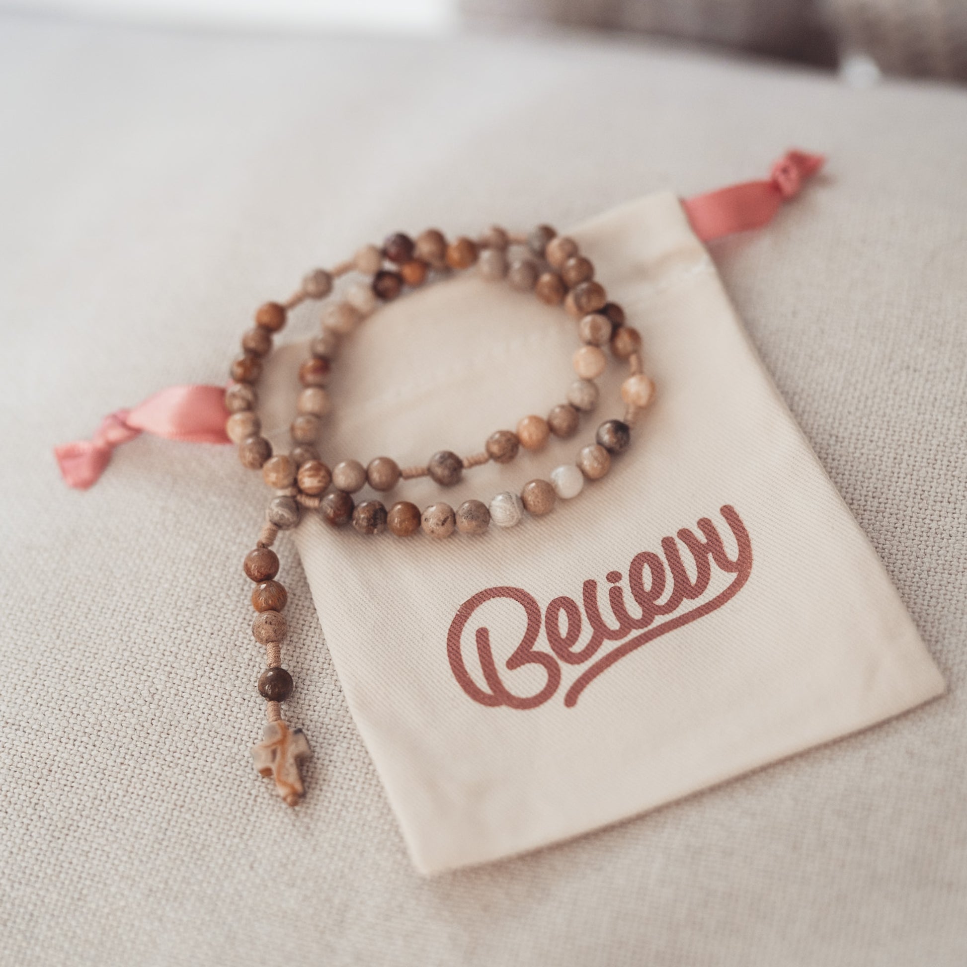 Różaniec z naturalnych kamieni w odcieniach brązu i beżu, zakończony krzyżykiem z kamienia, ułożony na beżowym woreczku z logo Believy i różową wstążką.