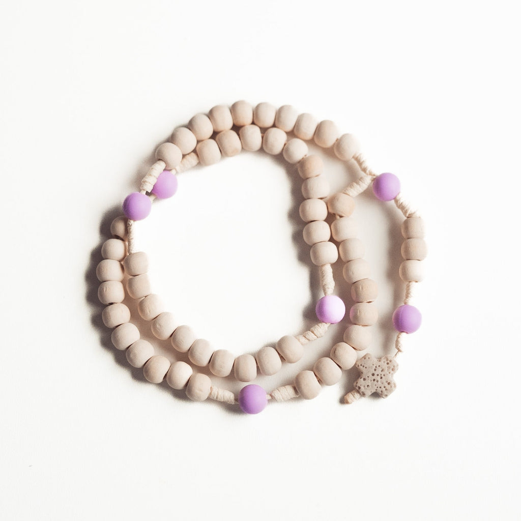 Delikatny różaniec z jasnych, drewnianych koralików przeplatanych fioletowymi kulkami i małym, beżowym krzyżykiem z koralika, ułożony na białym tle.