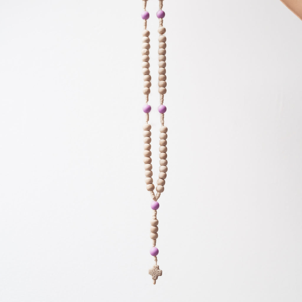 Różaniec z drewnianych, kremowych koralików i fioletowych akcentów, zakończony małym krzyżykiem, zawieszony na jasnym tle.