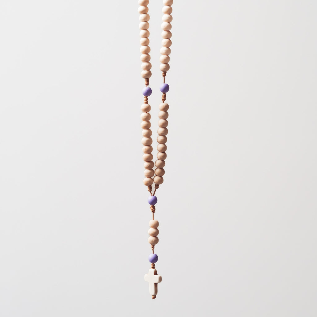 Drewniany różaniec z naturalnych, jasnych koralików i fioletowych akcentów, zawieszony na jasnym tle.