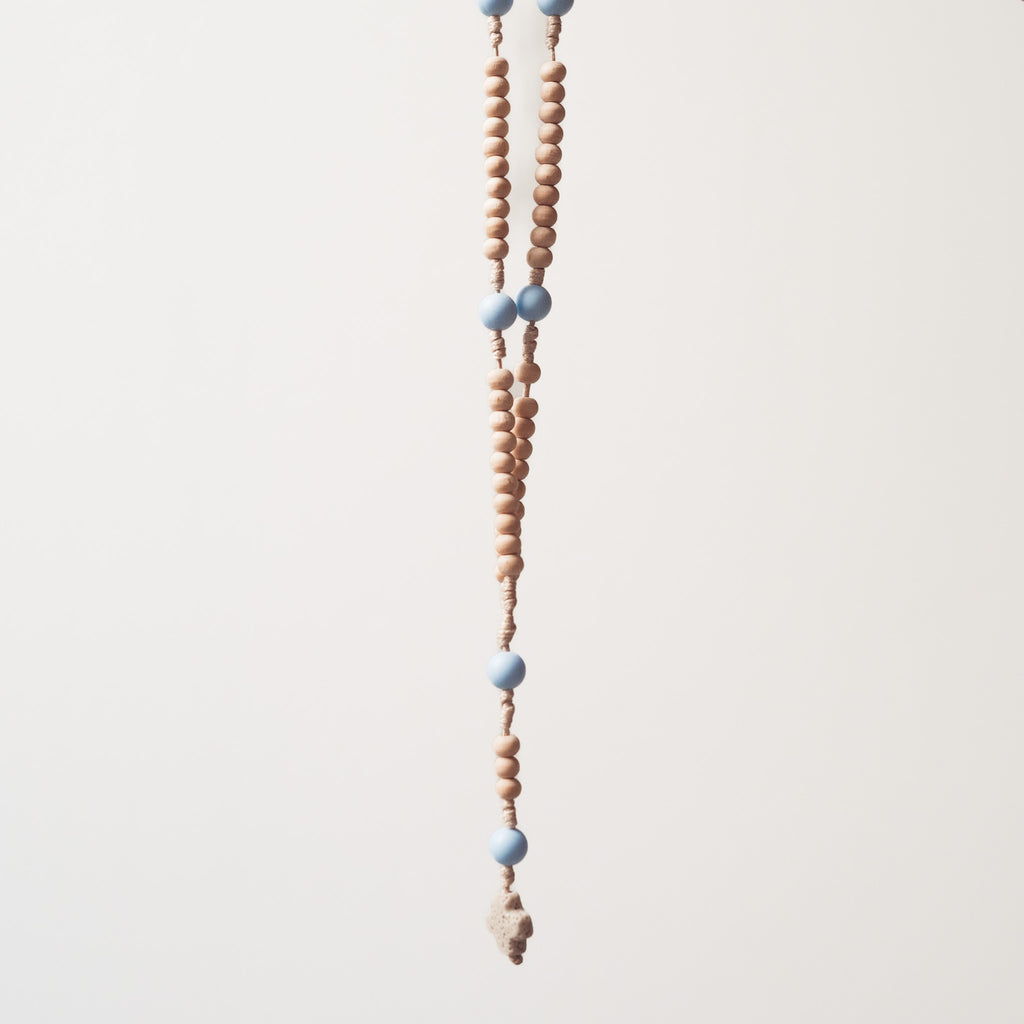 Drewniany różaniec z jasnymi koralikami i większymi, pastelowoniebieskimi akcentami, zawieszony na jasnym tle.