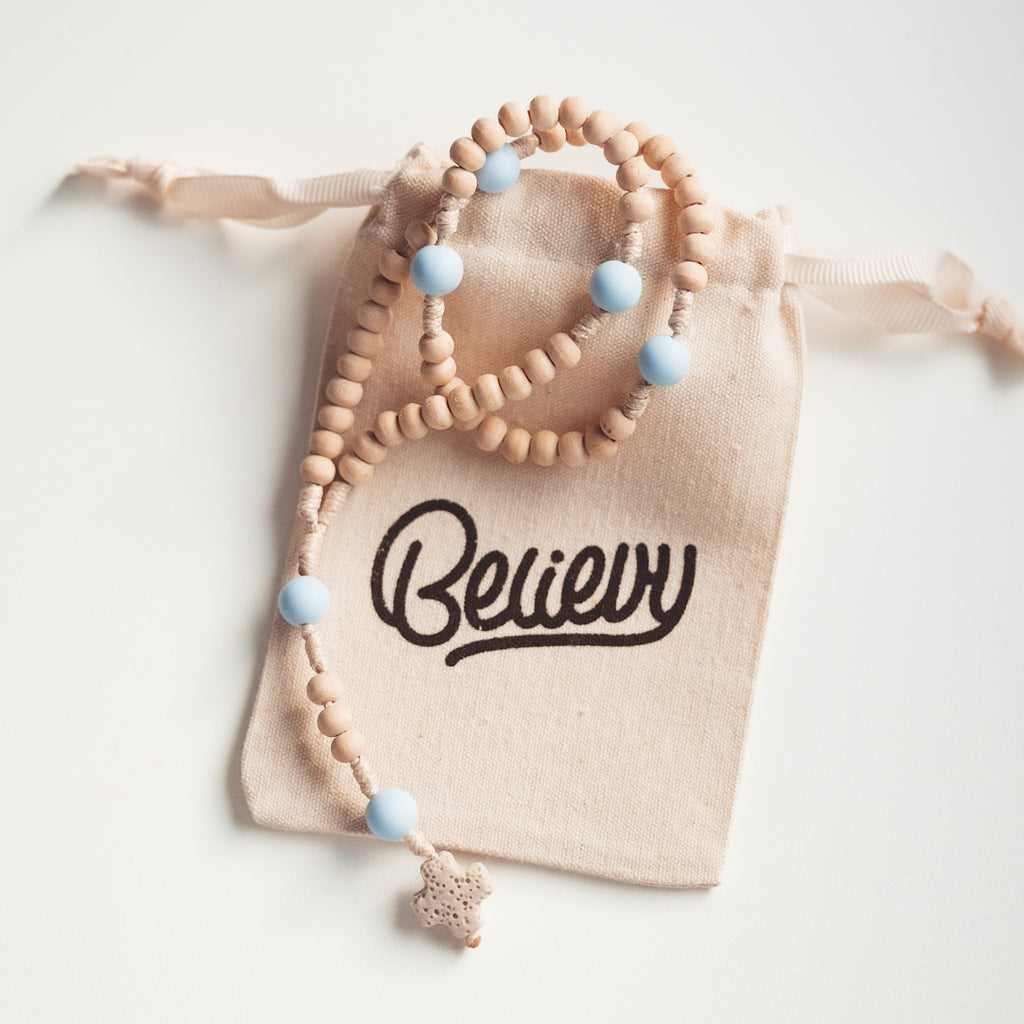 Różaniec z drewnianych koralików i niebieskich kulek, ułożony na beżowym woreczku z logo Believy.