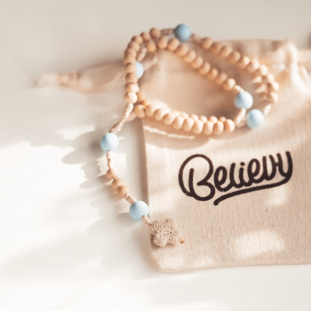 Drewniany różaniec z pastelowoniebieskimi koralikami, sfotografowany w delikatnym świetle, leżący na woreczku Believy.