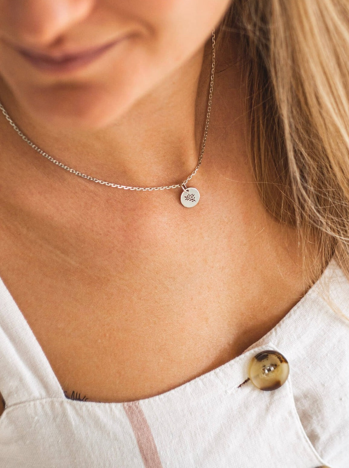 Srebrny naszyjnik - celebrytka na kobiecej szyi. Na łańcuszku widoczna okrągła zawieszka z wyciętym wzorem roślinnym - gałązką oliwną.