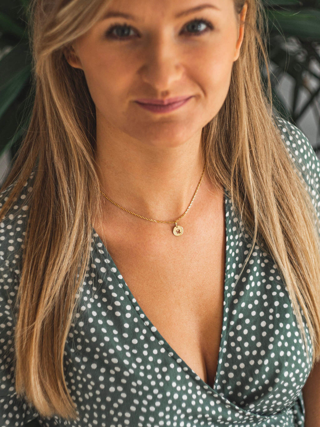 Uśmiechnięta modelka w zielonej sukience. Na jej szyi widnieje naszyjnik celebrytka z okrągłą zawieszką z wyciętym symbolem gołębia.  