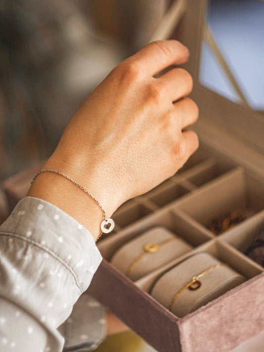 Kobieca ręka na szkatułką na biżuterię. Na jej nadgarstku widnieje srebrna bransoletka z okrągłą zawieszką z wyciętym symbolem serca.